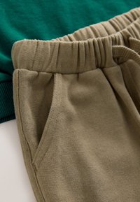 Gros plan d'un pantalon de survêtement beige à taille élastique avec poche latérale et tissu vert partiellement visible en dessous.