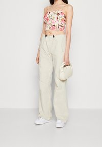 Kvinde iført hvide vidde bukser, hvide sneakers, blomsterfarvet pink crop top, mens hun holder en lille hvid håndtaske mod en ensfarvet baggrund.