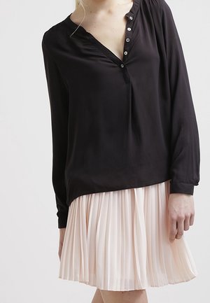 Blusa di seta nera con scollo a V e dettagli di bottoni, abbinata a una gonna plissettata rosa chiaro che arriva sopra il ginocchio.