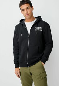 Jaunas vyras, dėvintis juodą zip-up gobtuvą su tekstu "Nirvana Nevermind" ir žaliomis krovinių kelnėmis, stovi su viena ranka kišenėje.