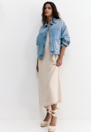 Ung kvinde iført en lyseblå denimjakke over en beige midi slip-kjole og cremefarvede espadrille kiler med ankelbånd, der står op ad en hvid væg.