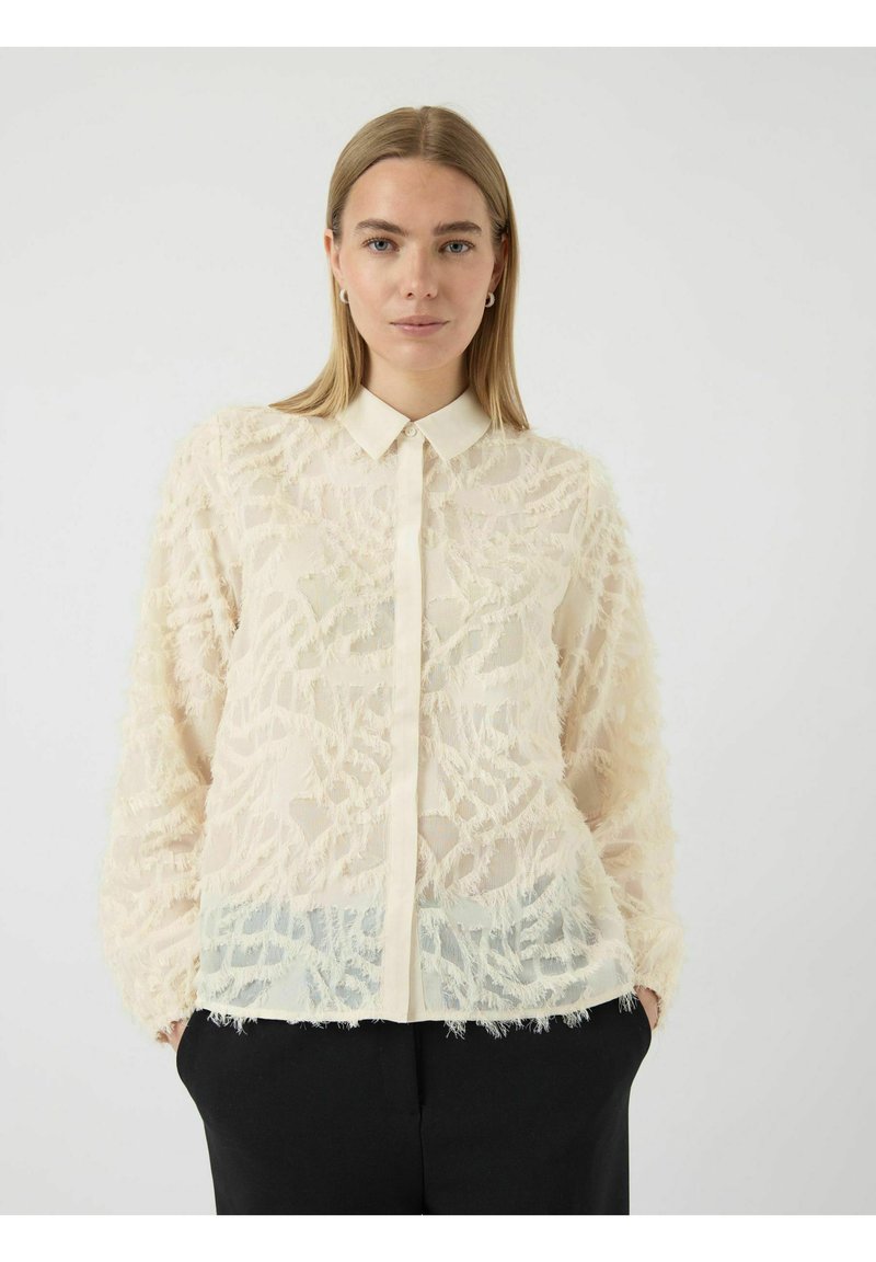 Frau mit glattem blonden Haar trägt eine cremefarbene, strukturierte, durchsichtige Bluse mit Knopfverschluss und schwarze Hosen, steht vor einem schlichten weißen Hintergrund.