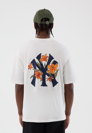 Uomo che indossa una maglietta bianca con un grande logo "NY" blu navy e gigli arancioni sul retro, abbinata a un berretto da baseball verde oliva e pantaloni neri.