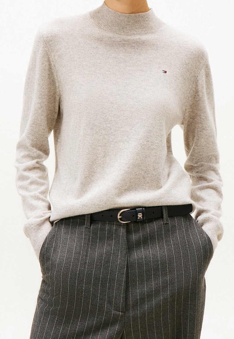 Pull long beige clair à manches longues avec petit logo, rentré dans un pantalon gris foncé à fines rayures avec ceinture noire et boucle dorée, mains dans les poches.