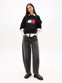 Pulôver preto de malha com um logótipo da Tommy Jeans em vermelho, branco e azul, combinado com jeans cinzentos de corte largo e mocassins pretos.