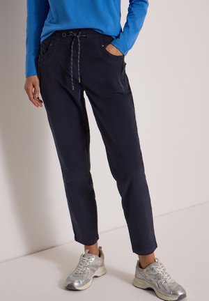 Personne portant un pantalon à cordon bleu marine, une chemise bleue à manches longues et des chaussures de sport argentées, debout devant un fond neutre.