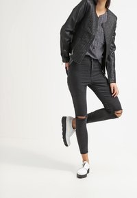 Svart läderjacka med quiltad yta, mönstrad skjorta, svarta rivna skinny jeans och vita skor med svarta accenter.