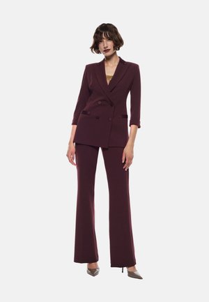 Blazer bordeaux doppiopetto con rever a punta, maniche a tre quarti, abbinato a pantaloni a gamba larga coordinati e décolleté a punta.