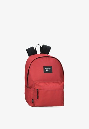 Mochila roja Reebok con correas negras, bolsillo frontal con cremallera y parche con el logotipo de Reebok en la parte superior delantera, mostrada sobre fondo blanco.