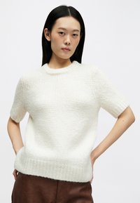 Pull à manches courtes en laine blanche douce et bouclée, avec un col rond et un ourlet côtelé, assorti à un pantalon brun texturé.