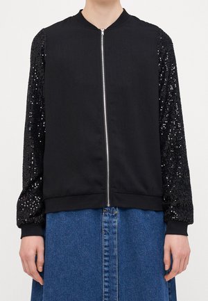 Veste noire zippée avec manches couvertes de sequins, portée sur une jupe en jean bleue, montrée du cou aux hanches.