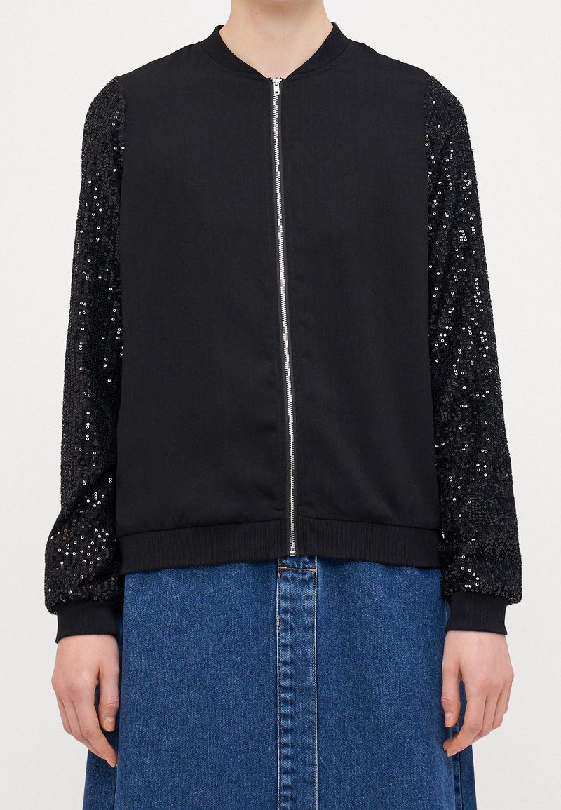 Veste noire zippée avec manches couvertes de sequins, portée sur une jupe en jean bleue, montrée du cou aux hanches.