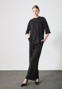 Svart oversized t-shirt med korta ärmar, kombinerad med svarta vida byxor och spetsiga klackar, mot en neutral bakgrund.