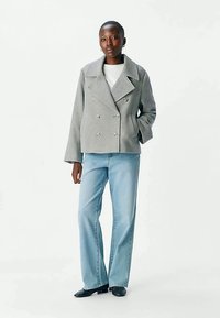 Kvinde står iført en grå dobbeltradet jakke, hvid top, lys blå vide jeans og sorte sko foran en ensfarvet lys baggrund.