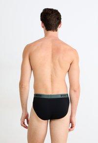 Modèle masculin de dos, portant des slips noirs avec une ceinture grise portant l'inscription "BOSS", devant un fond blanc uni.