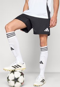 Crne sportske kratke hlače s bijelim naglascima, uparene s bijelim čarapama s crnim prugama. Bijele nogometne kopačke na uzorkovanoj lopti.