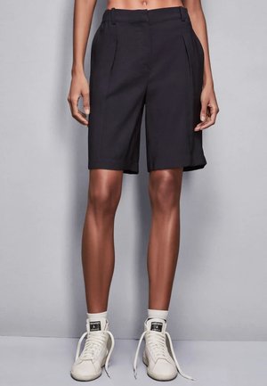 Pantaloni corti neri su misura con vita alta, pieghe e tasche laterali, abbinati a sneaker bianche e calze a costine. Tessuto liscio e design lunghezza ginocchio.