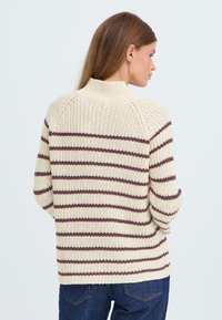 Dicker Strickpullover in Creme mit maroonfarbenen horizontalen Streifen. Besitzt einen hohen Kragen und eine gerippte Textur. Kombiniert mit dunkle blauen Jeans.