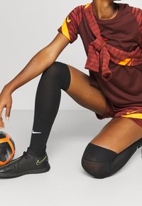 Nike Sportbekleidung in bordeauxrot mit orangefarbenen Akzenten; gemusterte Ärmel; knielange schwarze Socken; schwarze Schuhe mit gelben Details; Fußball.