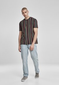 T-shirt noire avec des rayures verticales rouges, jaunes et vertes, associée à un jean bleu clair et des baskets grises, portée par un modèle sur un fond neutre.