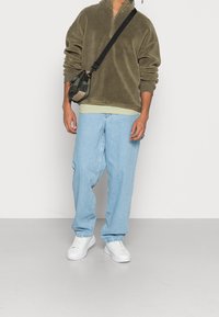 Felpa verde oliva con zip, strato leggero verde chiaro sotto, jeans azzurri larghi e sneakers bianche. Borsa a tracolla nera con motivo mimetico.