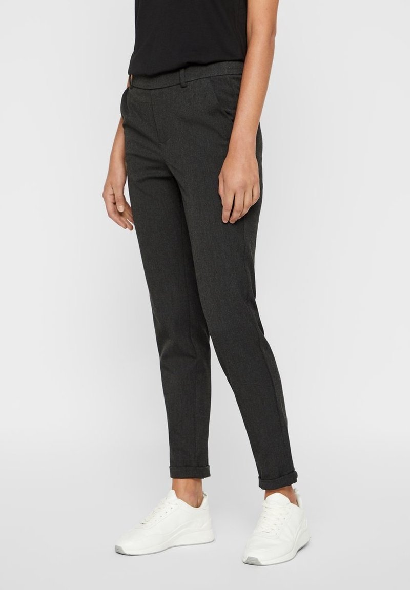 Personne portant un pantalon gris foncé fuselé avec des ourlets retroussés, un haut noir et des baskets blanches, debout devant un fond clair uni.