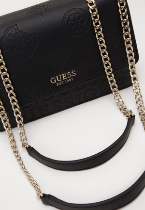 Sac Guess noir avec motifs de logo embossés, texte central doré "GUESS EST 1981", et bandoulière chaîne dorée avec épaulières en cuir noir.