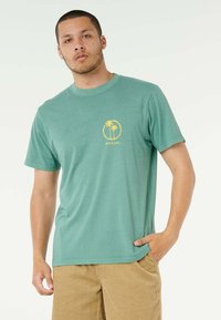 Giovane uomo che indossa una t-shirt verde Ripcurl con un logo di palma gialla, abbinata a pantaloncini in velluto a coste color sabbia, in piedi su uno sfondo bianco.