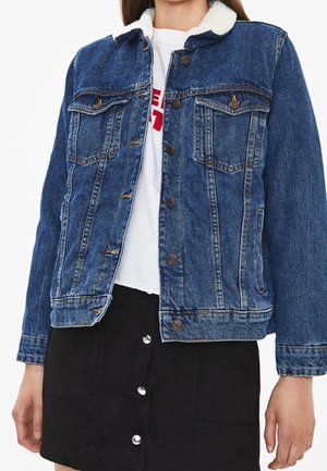 Femme portant une veste en jean bleue avec un col en polaire blanche par-dessus une chemise blanche et une jupe noire boutonnée.