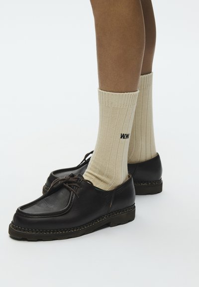 Persona valkā bēša krāsas ribotas zeķes ar melniem burtiem un melnas ādas kurpes ar šņorēm uz balta fona.