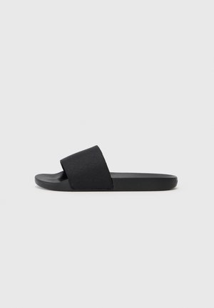 Sort slip-sandal med bred rem og flad sål, vist i profil på en ensfarvet lys grå baggrund.
