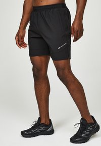 Homme portant un short de sport noir avec des poches latérales et des chaussures de trail noires, debout sur un fond clair uni.