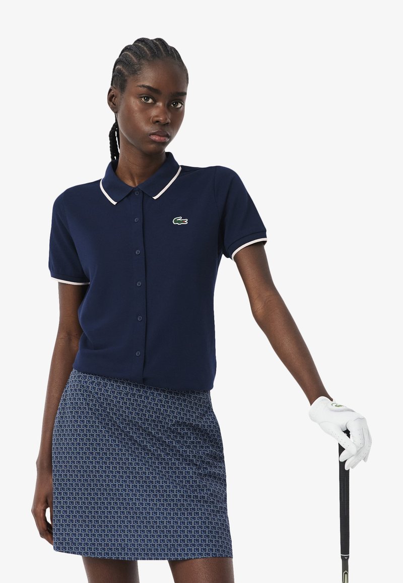 Frau trägt ein marineblaues Polohemd mit kurzen Ärmeln und einen gemusterten Rock, hält einen Golfclub mit weißem Handschuh an der rechten Hand.