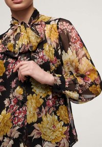 Blusa nera trasparente con motivo floreale giallo e rosa, dotata di collo legato, maniche lunghe e polsini con bottoni. Tessuto morbido con dettagli floreali.