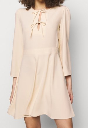 Femme portant une robe beige clair à manches longues avec deux nœuds noués au niveau de l'encolure et une jupe évasée.