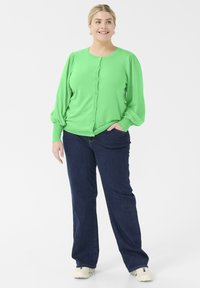 Grøn knap-opp cardigan med puffede ærmer, parret med mørkeblå jeans. Cardiganen har en glat tekstur og en rund halsudskæring.