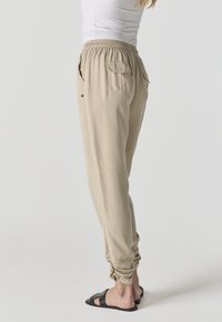 Femme portant un pantalon beige à taille élastique avec des poches à rabat latérales et arrière, associée à des sandales noires cloutées, debout sur un fond uni.