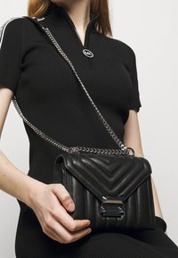 Mustan tikatun nahkalaukun, jossa on hopeinen ketjun kantohihna. Esittelee vinoraitakuviota ja metallista solkea sulkemista varten. Kompakti muotoilu.