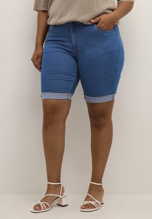 Person iført opsmøgede blå denimshorts, beige top og hvide sandaler med blokhæl og stropper, stående mod en ensfarvet baggrund.