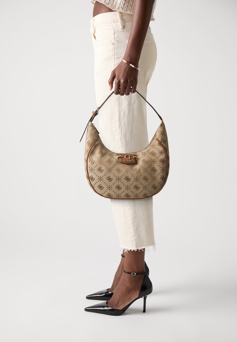 Femme tenant un sac à main Guess monogramme beige près d’un jean court crème et de talons aiguilles noirs à bride cheville sur fond blanc.