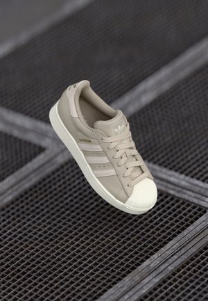 Beżowy sneaker Adidas Superstar z białym noskiem i trzema białymi paskami, umieszczony nad ciemną metalową kratą.