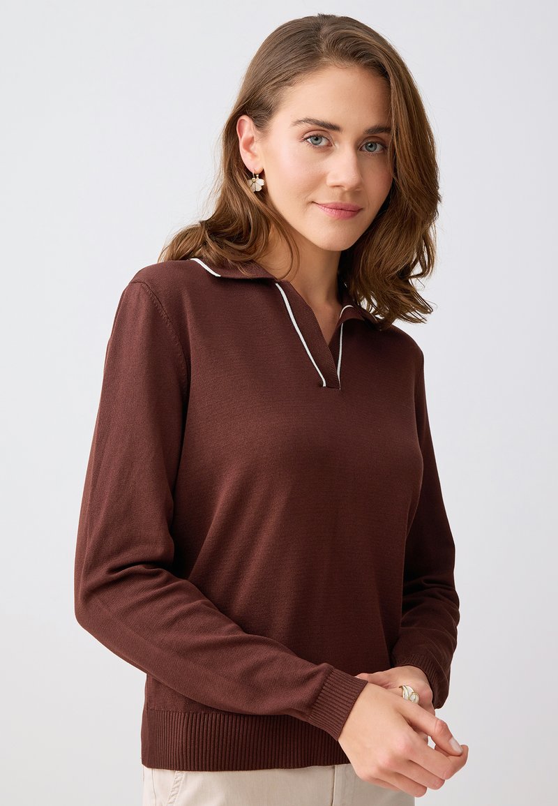 Femme aux cheveux bruns mi-longs portant un haut marron à manches longues avec un col bordé de blanc, un pantalon beige et des boucles d'oreilles en forme de fleur.