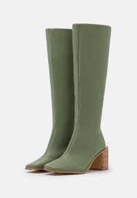 Botas hasta la rodilla de cuero verde con puntera cuadrada, tacón grueso con textura de madera a contraste y acabado liso. Detalle de cremallera a lo largo del lateral.
