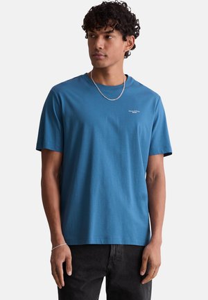 Mann mit lockigem Haar, der ein blaues kurzärmeliges T-Shirt und schwarze Hose trägt, schaut nach vorne und seitlich vor weißem Hintergrund.