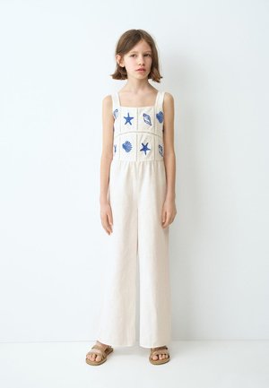 Mlada djevojka nosi bijeli kombinezon bez rukava s plavim uzorcima školjki i morskih zvijezda, stoji uz bijeli zid, nosi bež sandale.