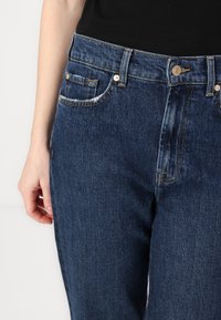 Jeans de mezclilla azul oscura con cintura de altura media, bolsillos frontales y costuras en contraste. Cuenta con un cierre de botón y cremallera.