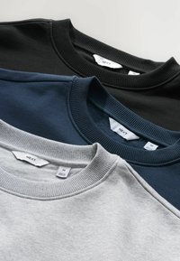 Tre foldede crewneck sweatshirt i sort, marineblå og lysegrå, hver med et hvidt NEXT mærke ved kraven.