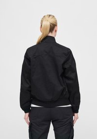 Schwarze Bomberjacke mit kragenförmigem Hals, gerippten Bündchen und Saum, glatter Textur und Rückennahtdetails, getragen über einem weißen Hemd.