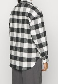 Long-sleeve shirt made of thick, checkered fabric in black and white, featuring a pointed collar and buttoned cuffs, with a rounded hemline.