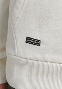Sudadera blanca con puños acanalados, que presenta una superficie texturizada y una etiqueta negra con texto blanco. El diseño muestra un detalle mínimo.